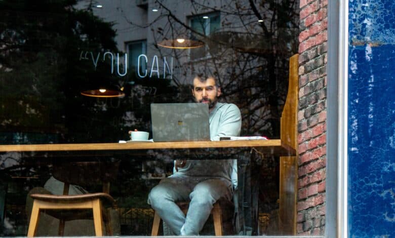 Ein Mann sitzt in einem Café vor seinem Laptop mit einer Tasse Kaffee und sieht aus dem Fenster.