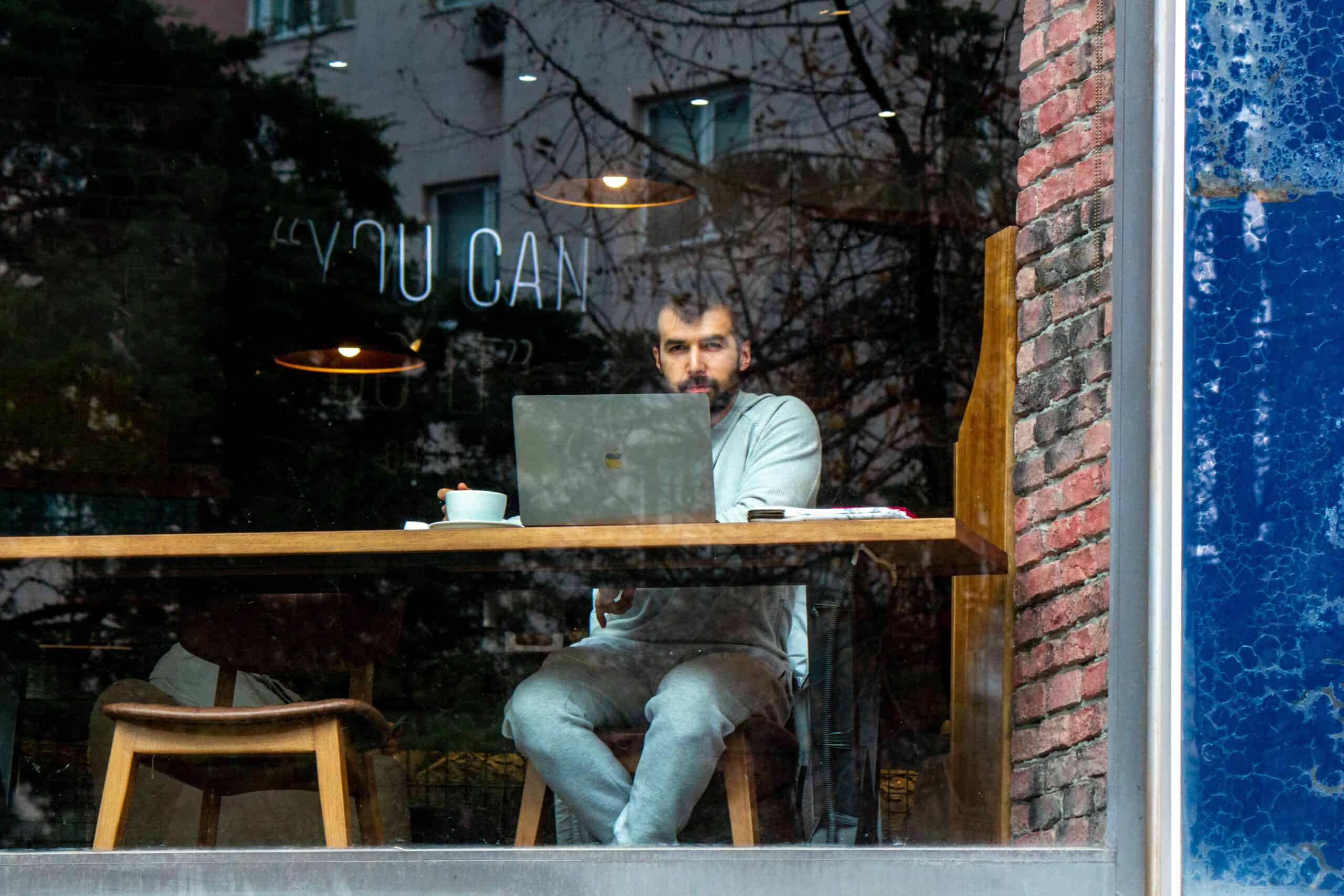 Ein Mann sitzt in einem Café vor seinem Laptop mit einer Tasse Kaffee und sieht aus dem Fenster.